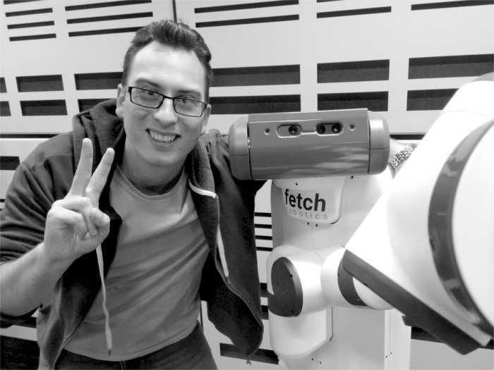 Andrew Haigh smiles at the camera, throwing a peace sign in his
	right hand and with his left hand around the shoulder of a Fetch Robotics
	mobile manipulator